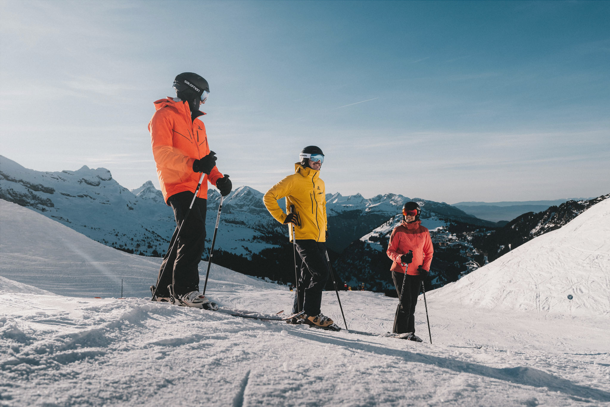 Three skiers in colourful outfits at a standstill on a snow-covered slope, with the Flaine ski area and the Aravis mountains in the background under a clear sky.