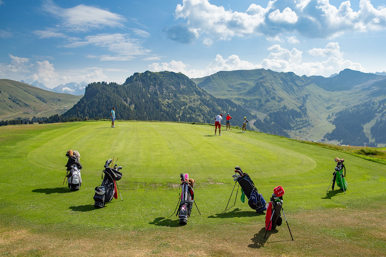 Des golfeurs s'exercent sur le parcours de Flaine, un green verdoyant qui offre une vue spectaculaire sur les massifs montagneux environnants. Au premier plan, plusieurs sacs de golf colorés sont posés sur l'herbe sous un ciel bleu parsemé de nuages.